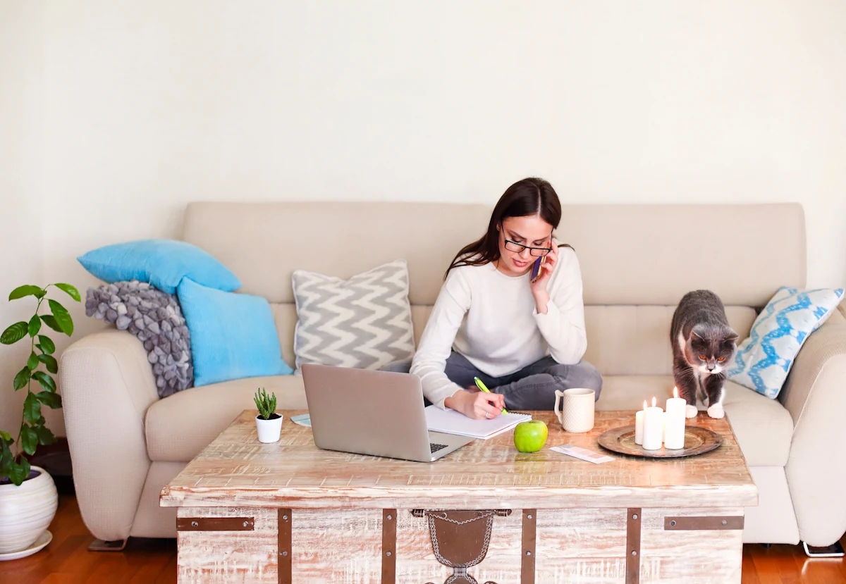Une femme au téléphone assise dans son canapé et devant son ordinateur entrain de prendre des notes